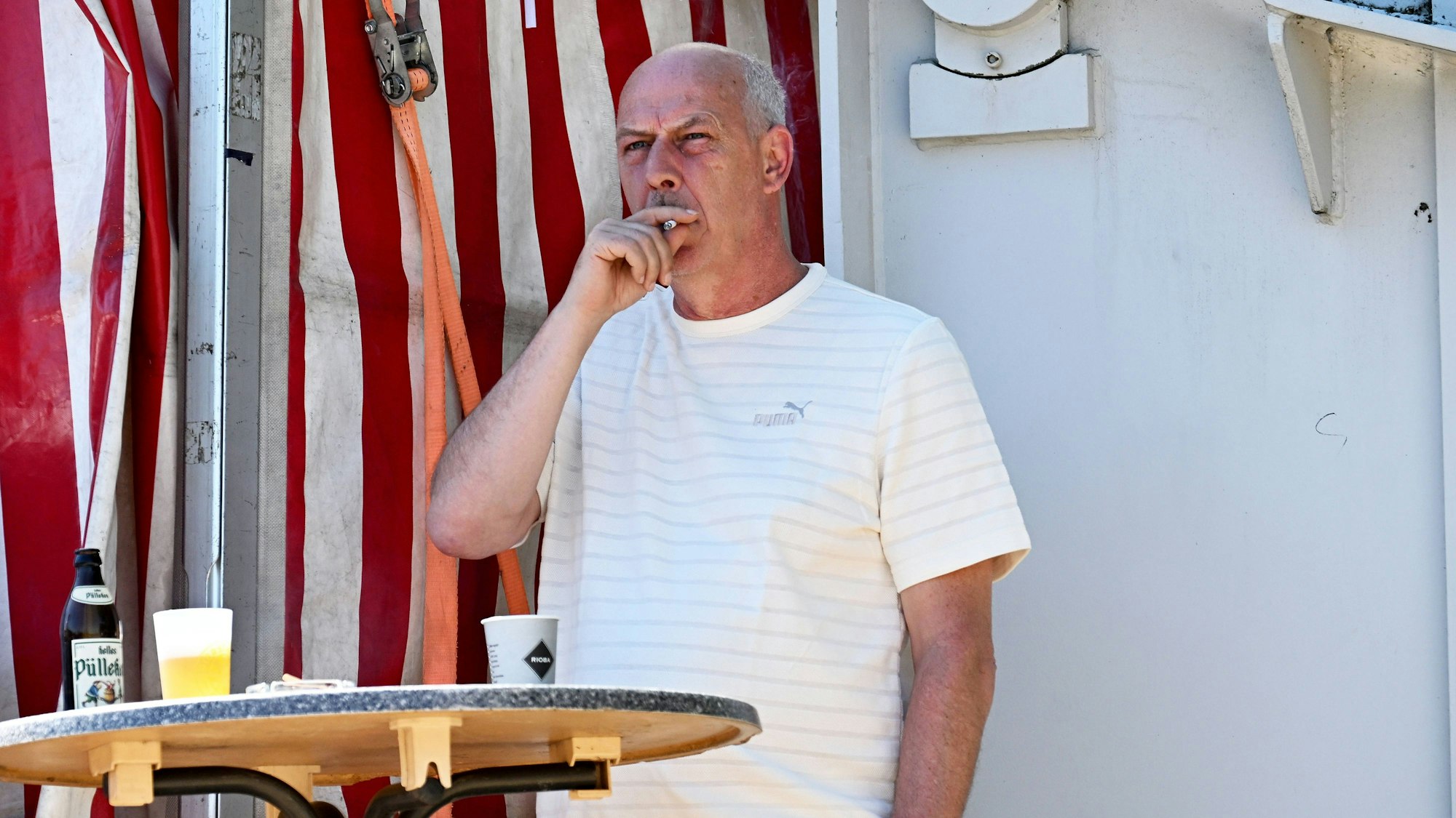 Mario Basler raucht an einem Stehtisch eine Zigarette. Auf dem Tisch stehen ein Kaffeebecher und eine Flasche Pils.