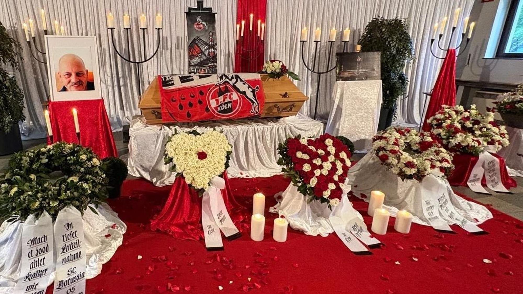 Auf einem Sarg liegt die Fahne des 1. FC Köln, daneben ein Foto des Toten, auch die Blumen sind in rot-weiß gehalten.