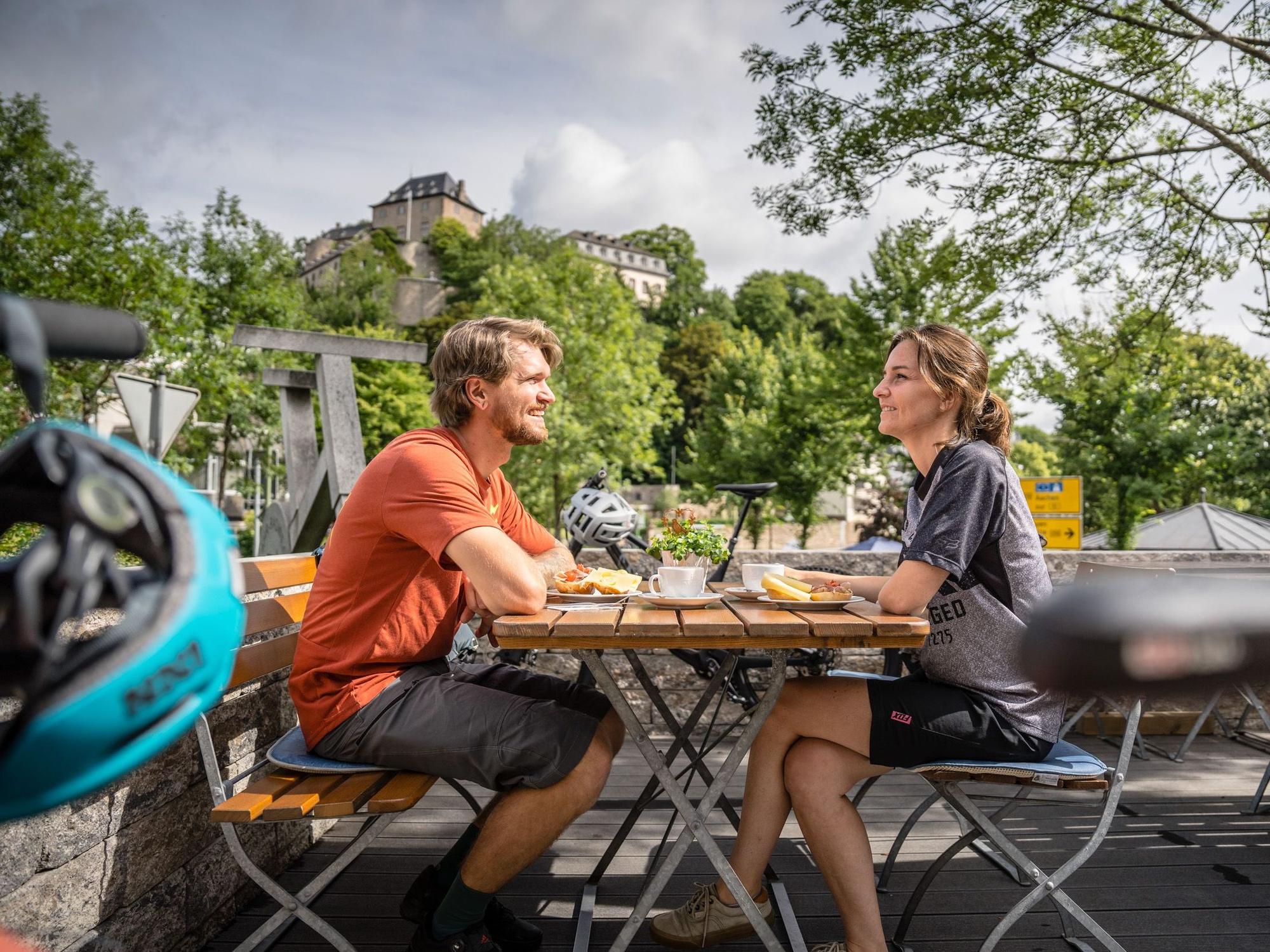 Zwei Radfahrer machen eine Pause und sitzen an einem Biergartentisch. Im Hintergrund auf einem Berg eine Burg.