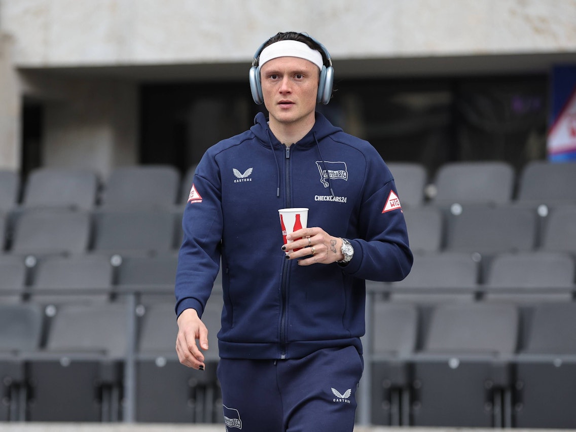 Fabian Reese betritt mit Trainingsanzug, Kopfhörern und einem Trinkbecher den Platz.