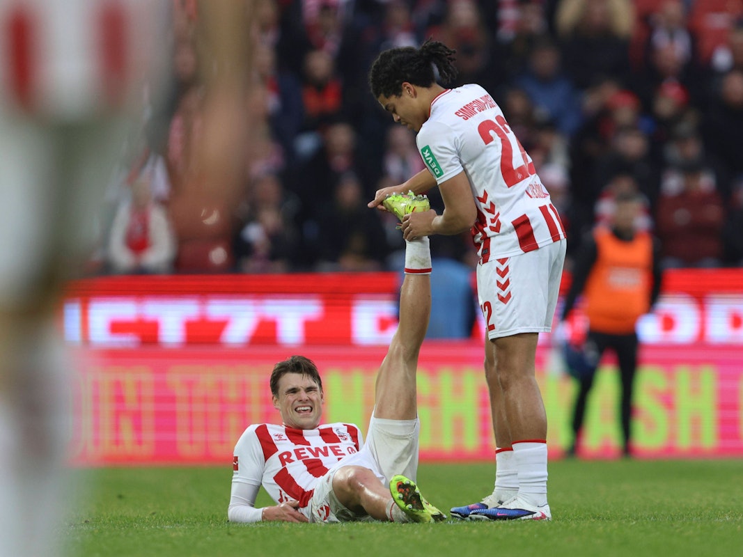 Joel Schmied (1. FC Köln) liegt verletzt am Boden, Jahmai Simpson-Pusey dehnt sein Bein.