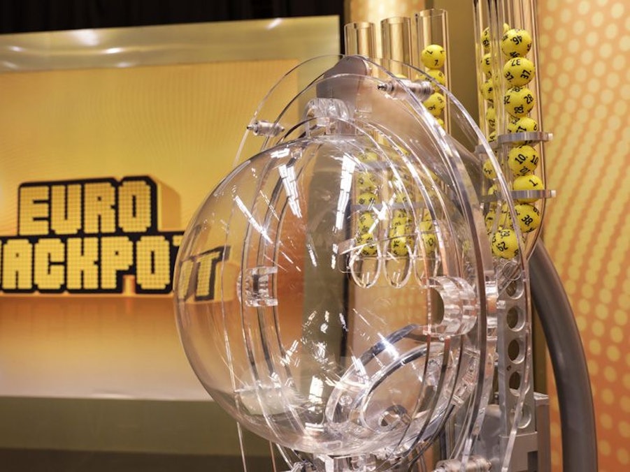 Das Bild zeigt die moderne Ziehungsmaschine für die Eurojackpot-Lotterie. Im Hintergrund ist groß das goldene „EURO JACKPOT“-Logo auf einer beleuchteten Wand zu sehen.