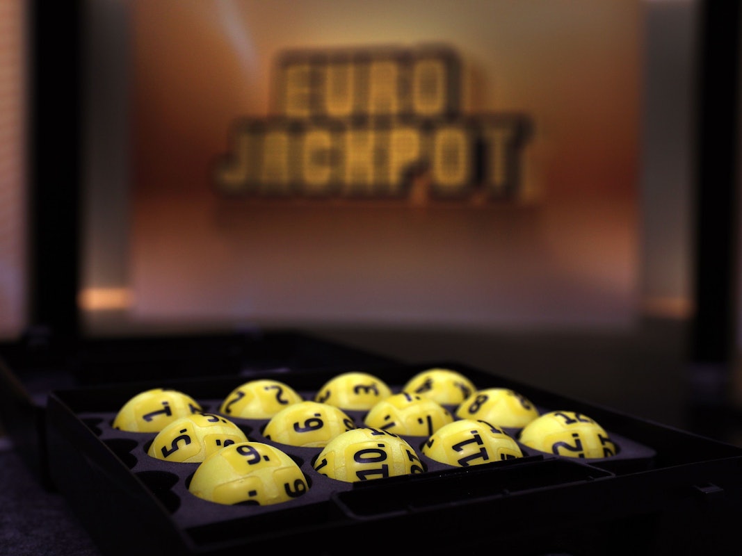 Das Bild zeigt mehrere gelbe Lotteriekugeln mit schwarzen Nummern, die ordentlich in einem schwarzen Tablett angeordnet sind. Im unscharfen Hintergrund ist das beleuchtete Eurojackpot-Logo zu erkennen.