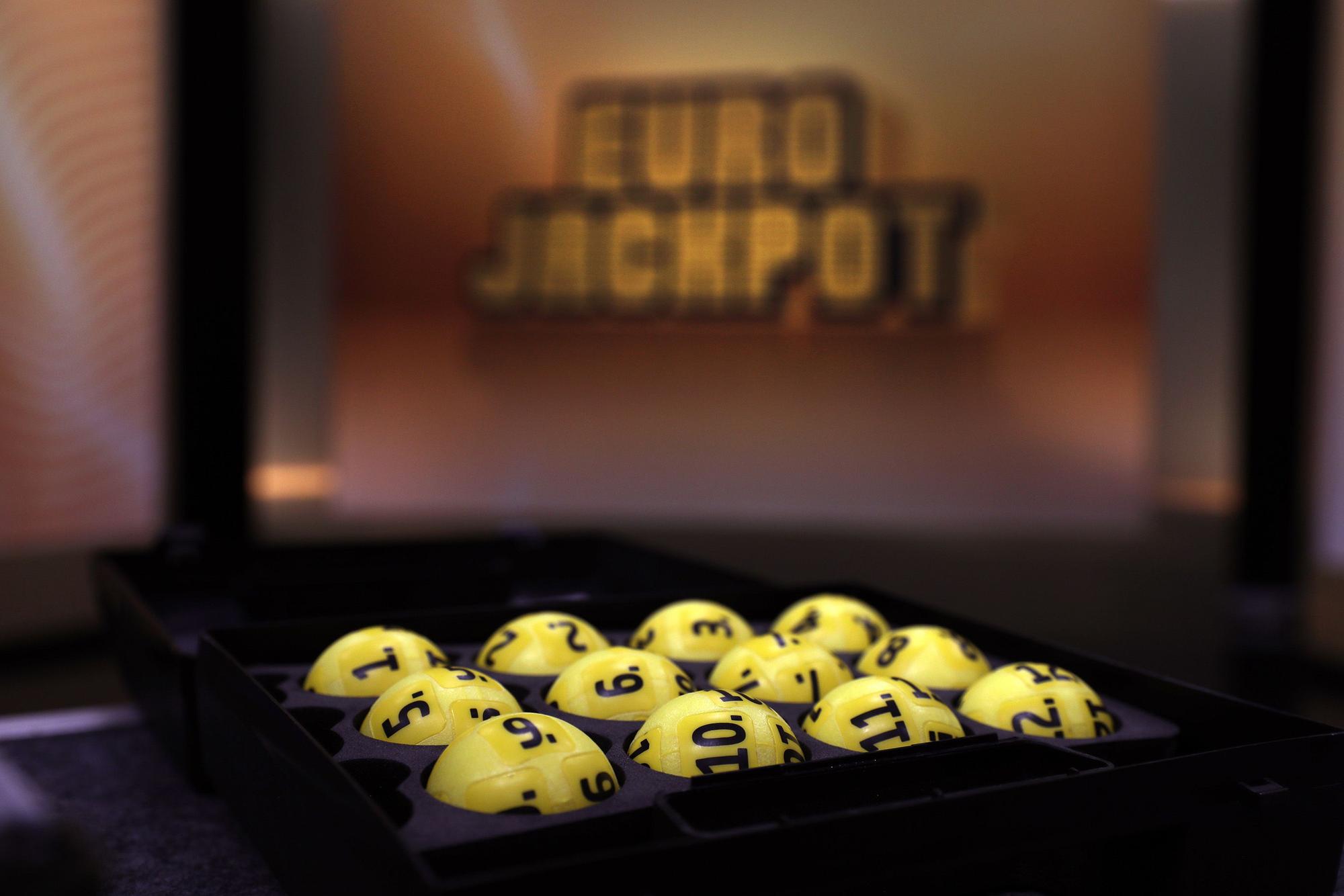 Das Bild zeigt mehrere gelbe Lotteriekugeln mit schwarzen Nummern, die ordentlich in einem schwarzen Tablett angeordnet sind. Im unscharfen Hintergrund ist das beleuchtete Eurojackpot-Logo zu erkennen.