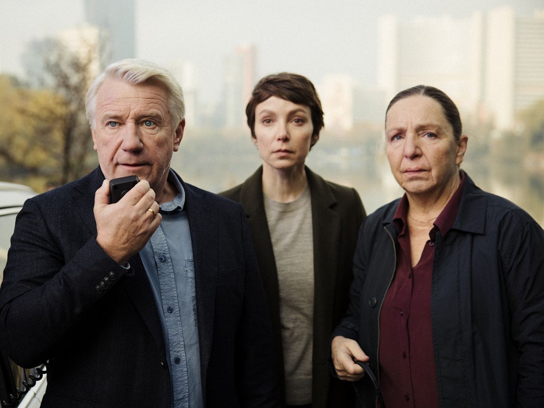 Ein Mann mit grauen Haaren spricht in ein Diktiergerät, neben ihm stehen zwei Frauen und schauen gebannt zu