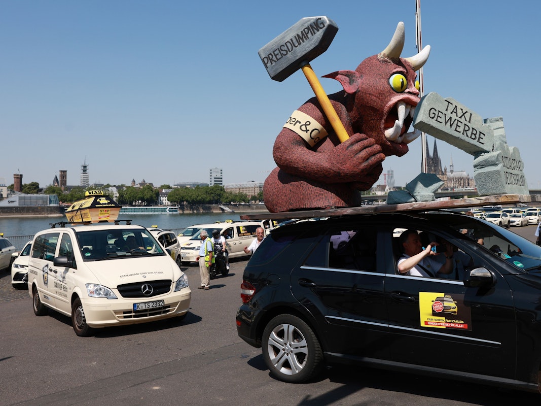 Auf einem Autodach ist eine Pappfigur mit Hörnern, die einen Hammer schwingt. Hinter dem Wagen stehen Taxis Schlange.
