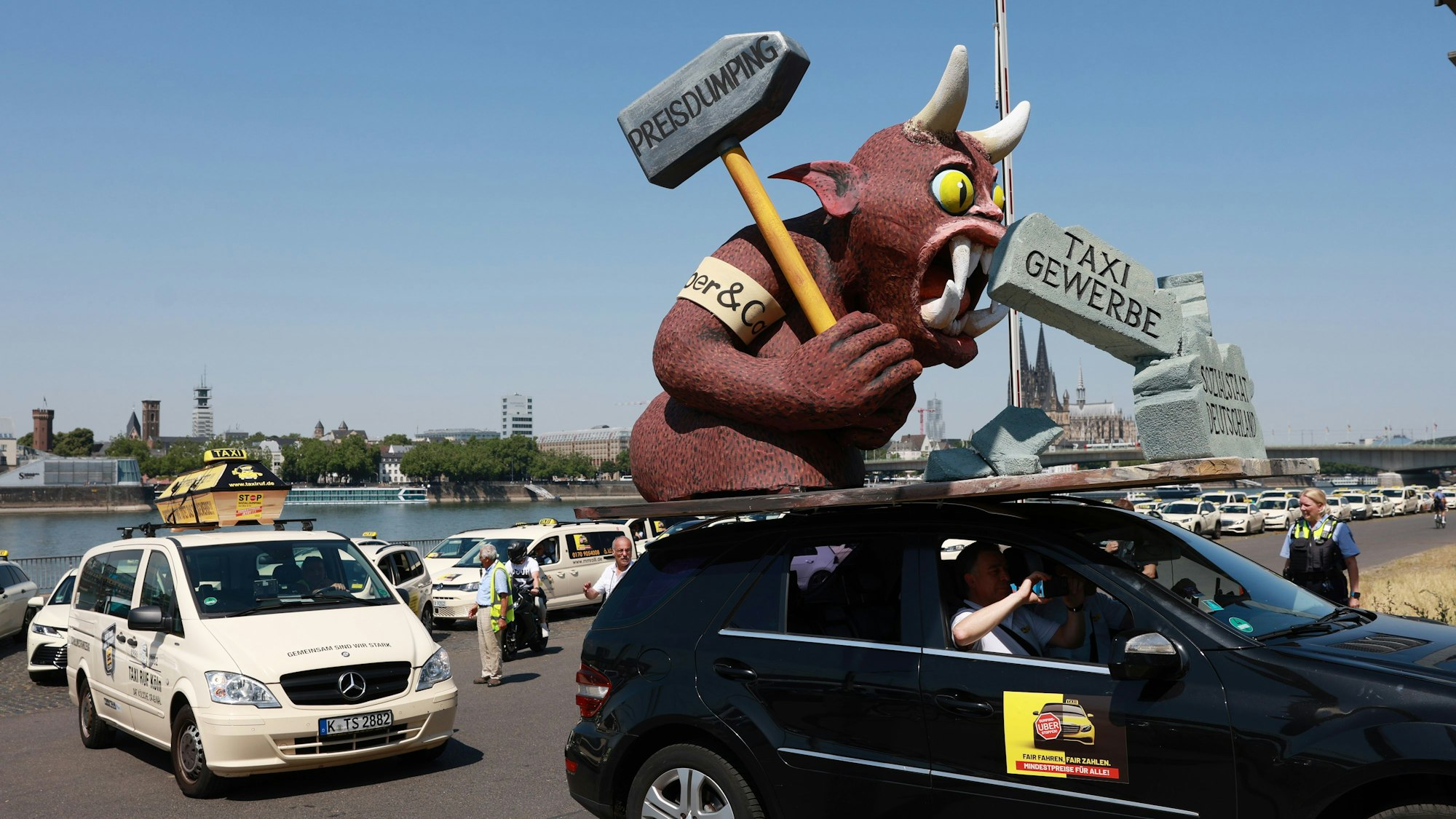Auf einem Autodach ist eine Pappfigur mit Hörnern, die einen Hammer schwingt. Hinter dem Wagen stehen Taxis Schlange.