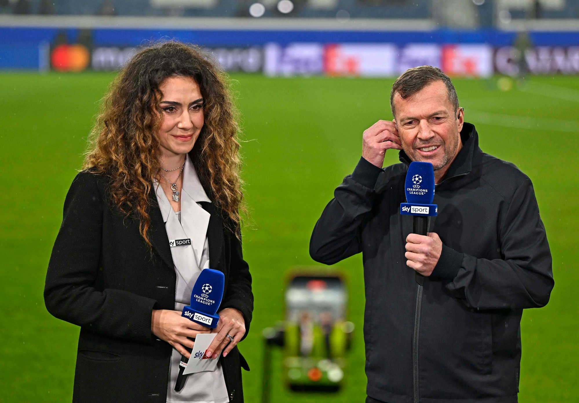 Lothar Matthäus an der Seite von Kimberly Budinsky mit Mikros in der Hand auf dem Rasen.