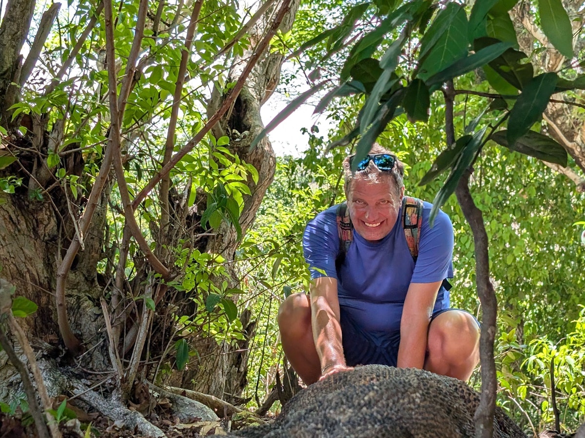 Ein Mann hockt hinter einer riesigen Echse im Dschungel und lacht.