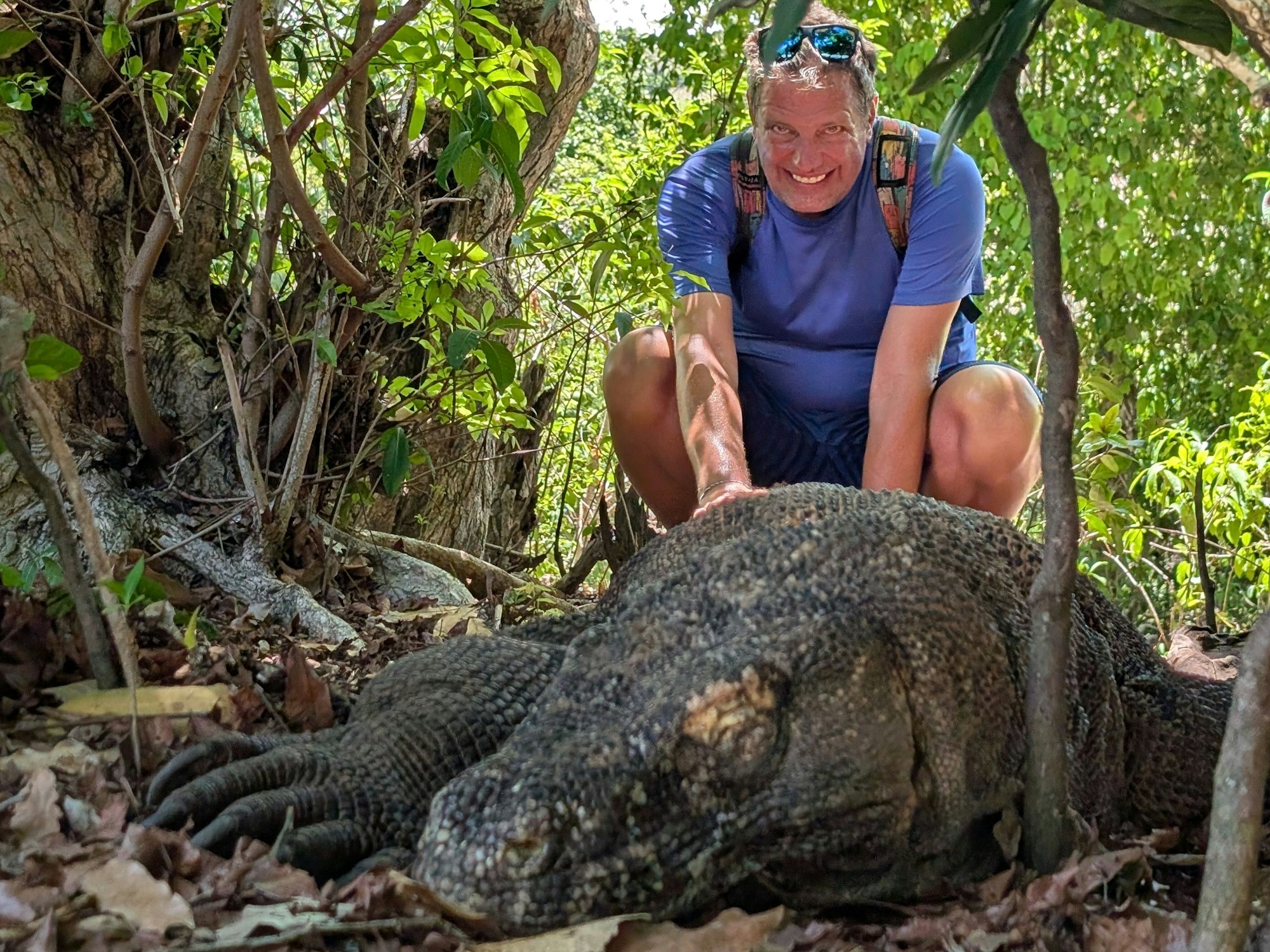 Ein Mann hockt hinter einer riesigen Echse im Dschungel und lacht.