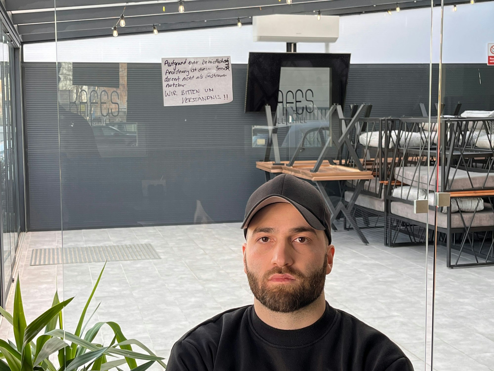 Kerim Akpinar steht vor einer überdachten Terrasse, auf der Stühle und Tische zusammengestapelt wurden.