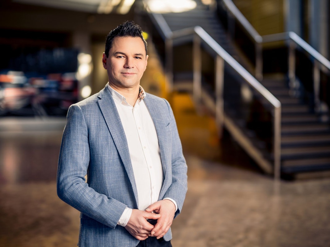 Ein Mann in Anzug und weißem Hemd hat die Hände zur Raute gefaltet und lächelt in die Kamera. Im Hintergrund ist eine Treppe zu sehen.