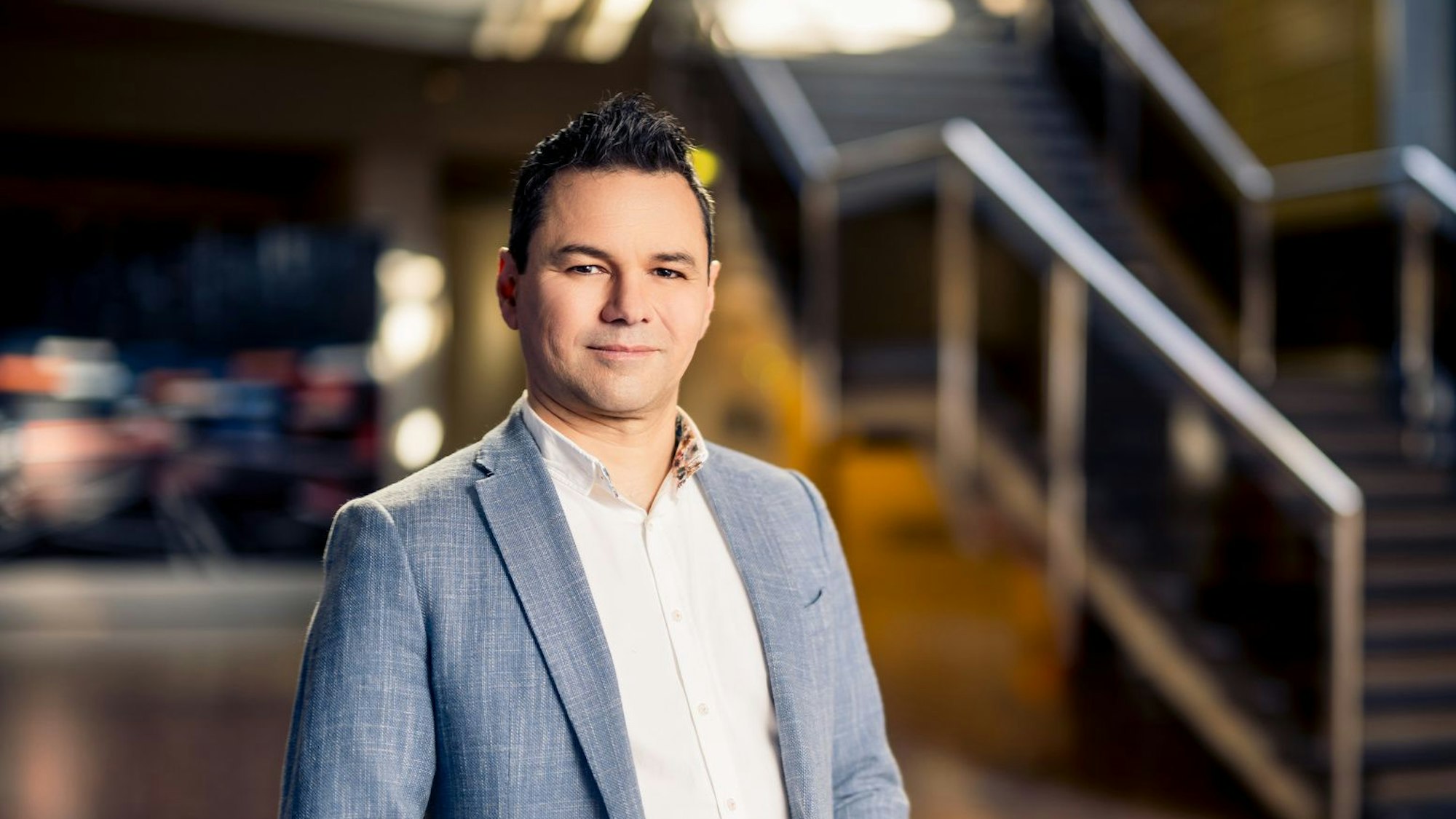 Ein Mann in Anzug und weißem Hemd hat die Hände zur Raute gefaltet und lächelt in die Kamera. Im Hintergrund ist eine Treppe zu sehen.