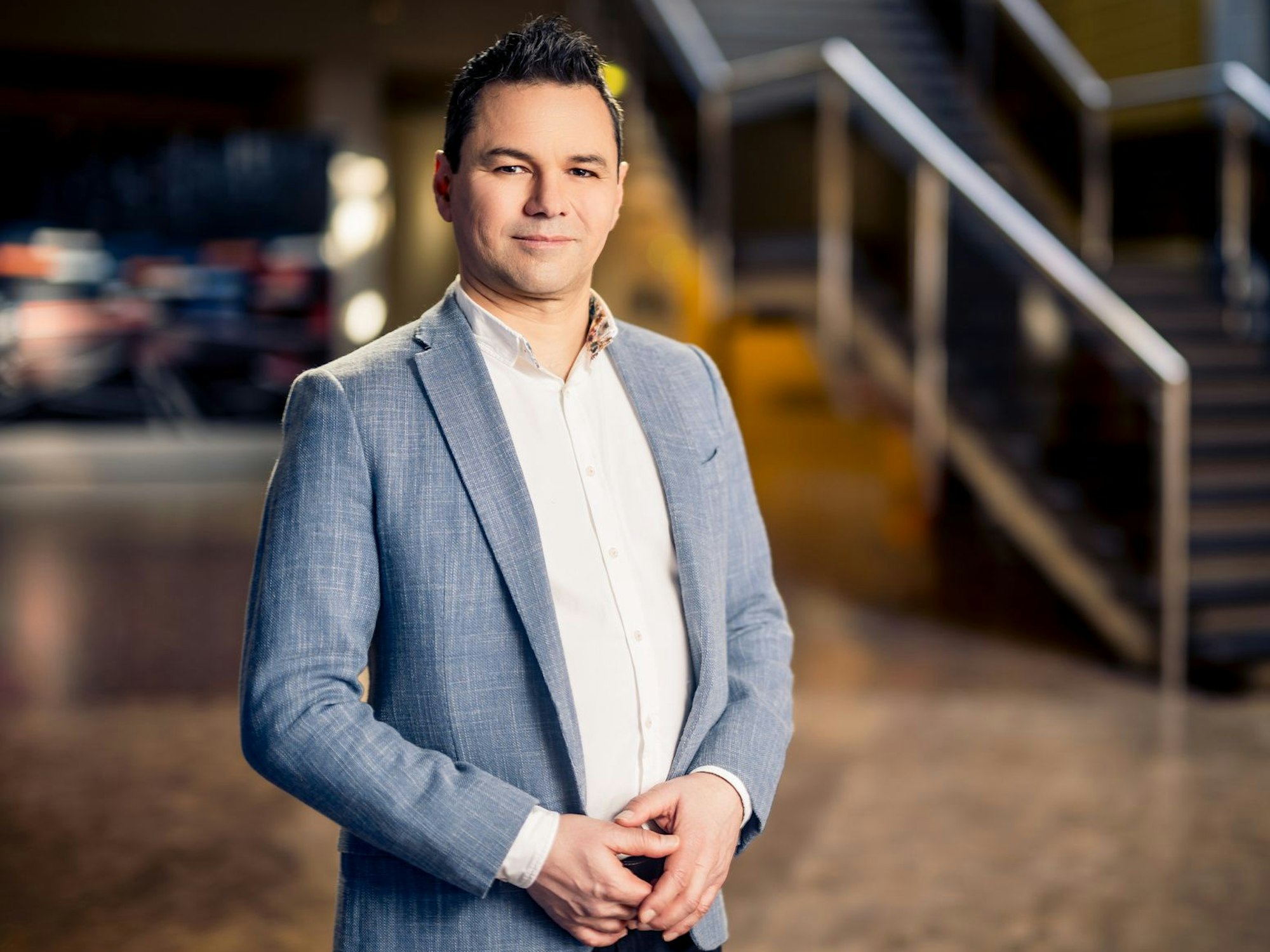 Ein Mann in Anzug und weißem Hemd hat die Hände zur Raute gefaltet und lächelt in die Kamera. Im Hintergrund ist eine Treppe zu sehen.