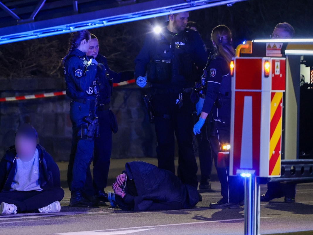19.03.2026, Nordrhein-Westfalen, Mönchengladbach: Zwei Tatverdächtige sitzen und liegen in Handschellen gefesselt auf einer Straße, bewacht von Polizeibeamten. In der Innenstadt waren zuvor Schüsse gefallen. Es gebe Verletzte, sagte ein Polizeisprecher. Foto: Christoph Reichwein/dpa - ACHTUNG: Tatverdächtige Person wurde aus rechtlichen Gründen gepixelt +++ dpa-Bildfunk +++