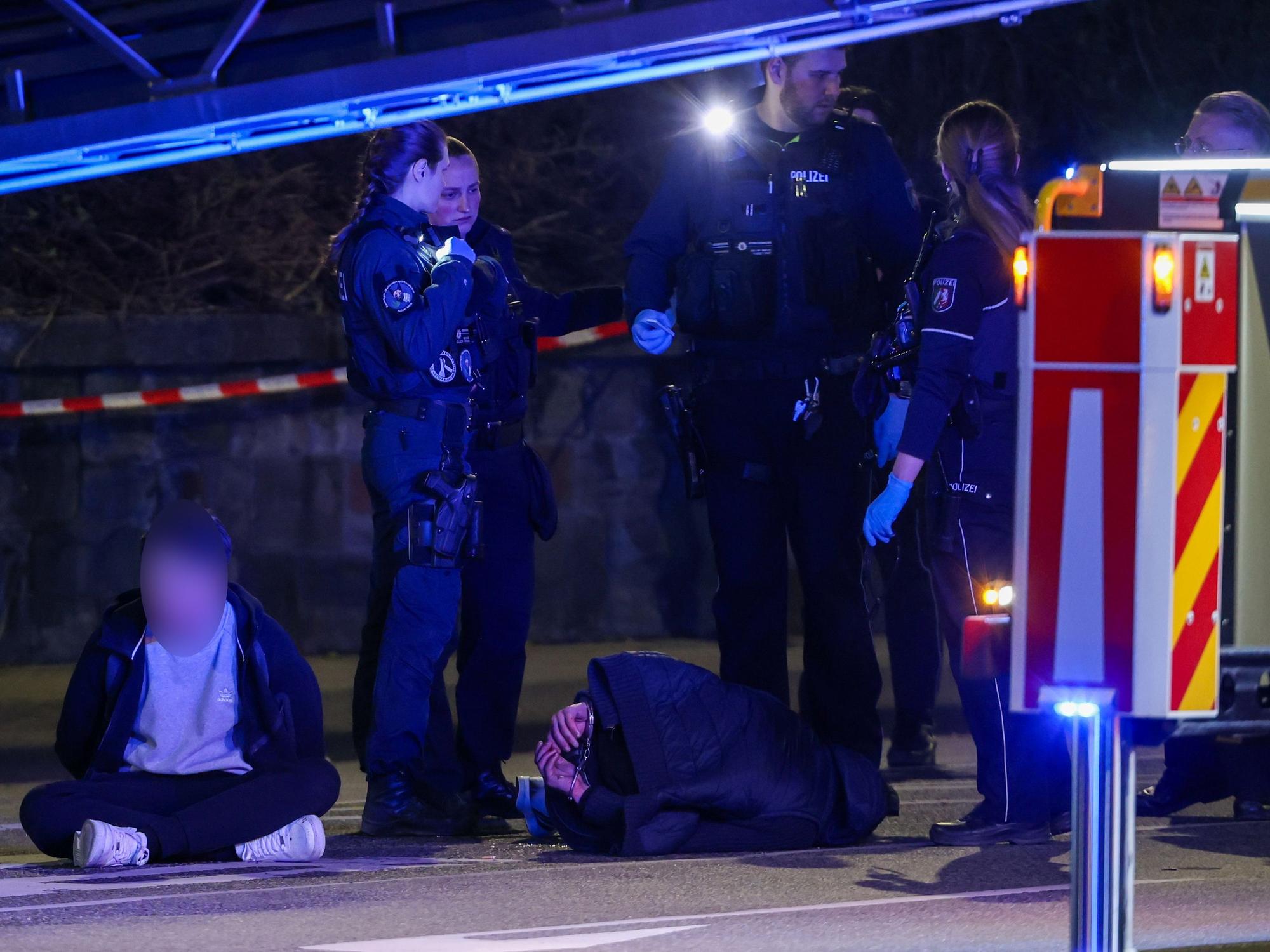 19.03.2026, Nordrhein-Westfalen, Mönchengladbach: Zwei Tatverdächtige sitzen und liegen in Handschellen gefesselt auf einer Straße, bewacht von Polizeibeamten. In der Innenstadt waren zuvor Schüsse gefallen. Es gebe Verletzte, sagte ein Polizeisprecher. Foto: Christoph Reichwein/dpa - ACHTUNG: Tatverdächtige Person wurde aus rechtlichen Gründen gepixelt +++ dpa-Bildfunk +++
