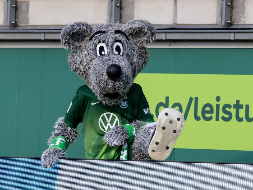 Maskottchen Wölfi vom VfL Wolfsburg klettert über eine Werbebande.