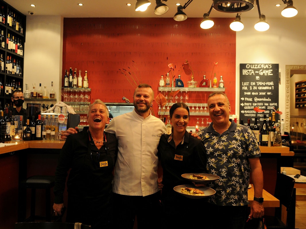 Zwei Frauen und zwei Männer stehen an der Theke eines Restaurants, eine der Frauen hält zwei angerichtete Teller in der Hand.