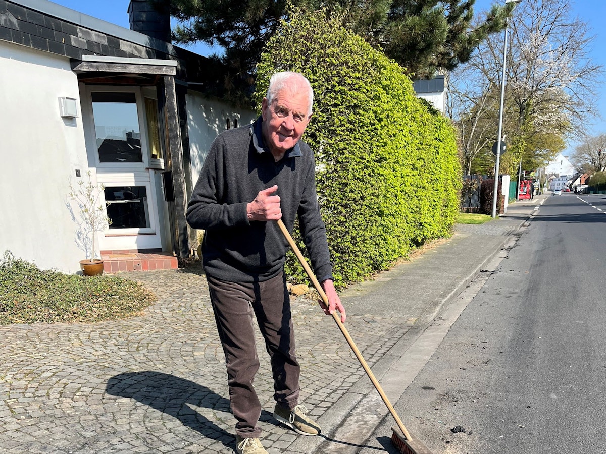 Ein älterer Mann steht mit einem Besen an der Straße.