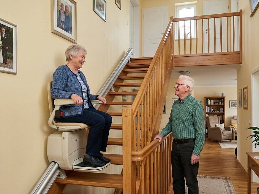 Eine Seniorin sitzt auf einem Treppenlift und fährt die Treppe eines Hauses hoch.