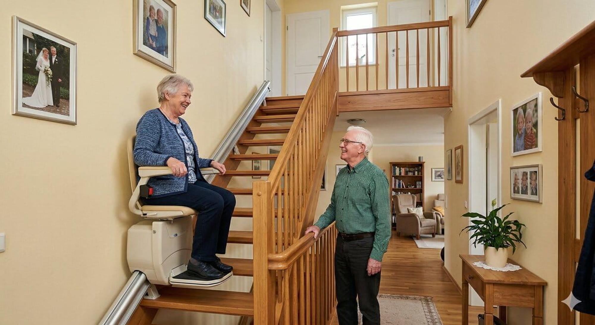 Eine Seniorin sitzt auf einem Treppenlift und fährt die Treppe eines Hauses hoch.