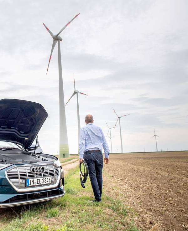 Ein Mann mit Ladekabel steht neben einem E-Auto im Windpark