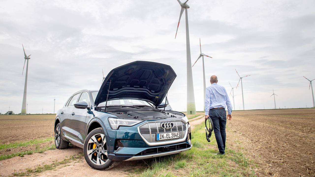 Ein Mann mit Ladekabel steht neben einem E-Auto im Windpark