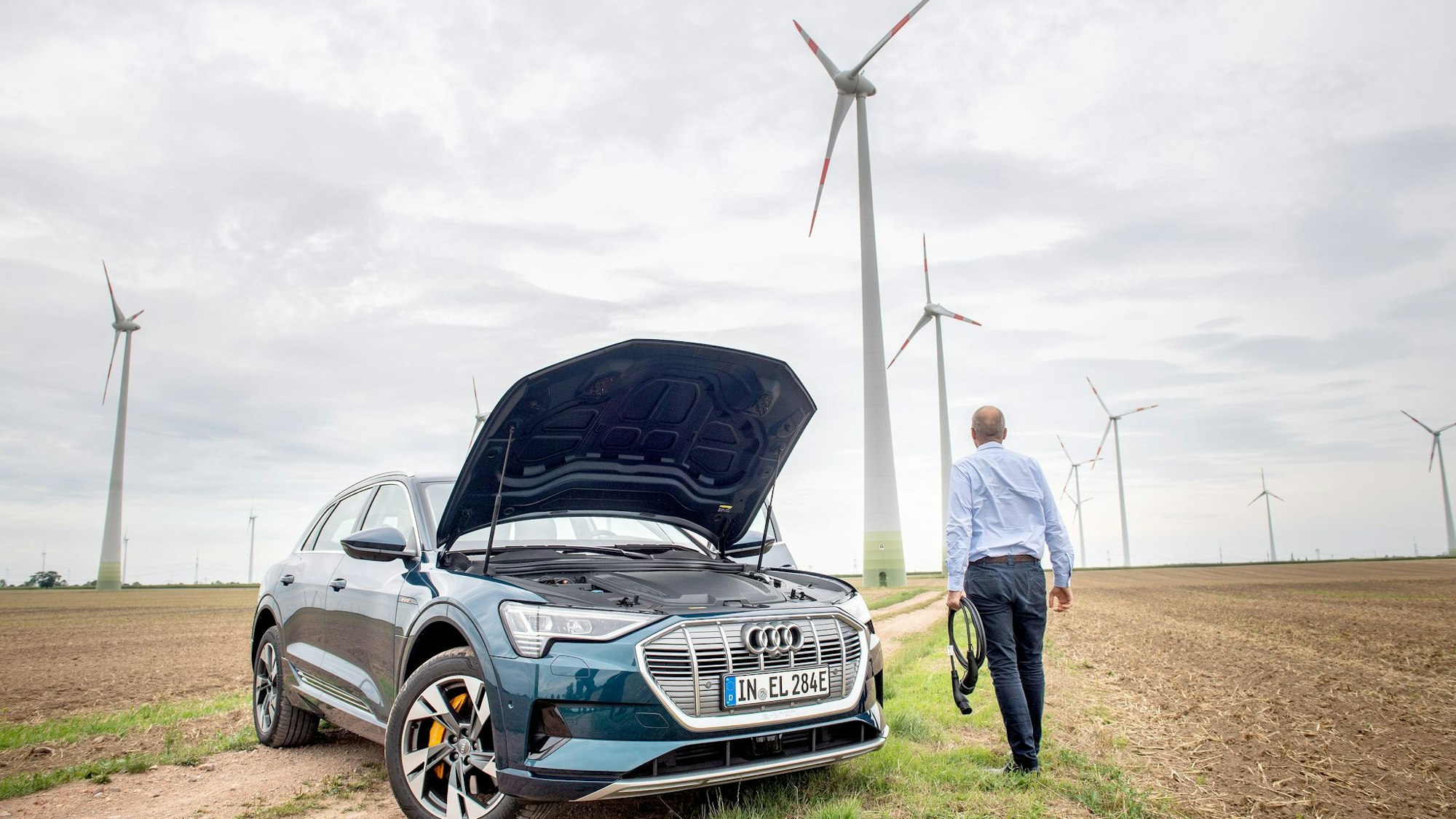 Ein Mann mit Ladekabel steht neben einem E-Auto im Windpark