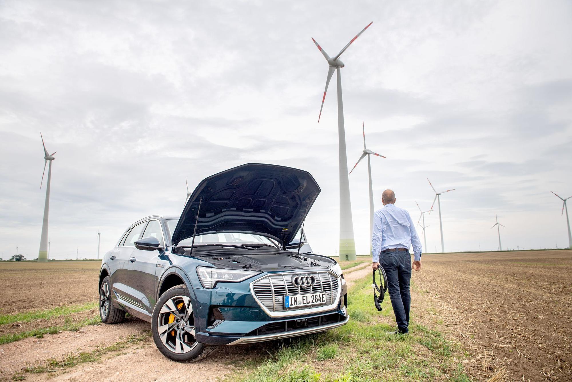 Ein Mann mit Ladekabel steht neben einem E-Auto im Windpark