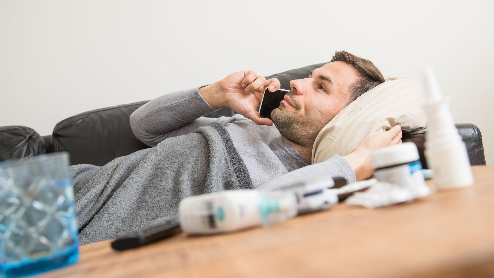 Ein Mann liegt krank auf einem Sofa und telefoniert