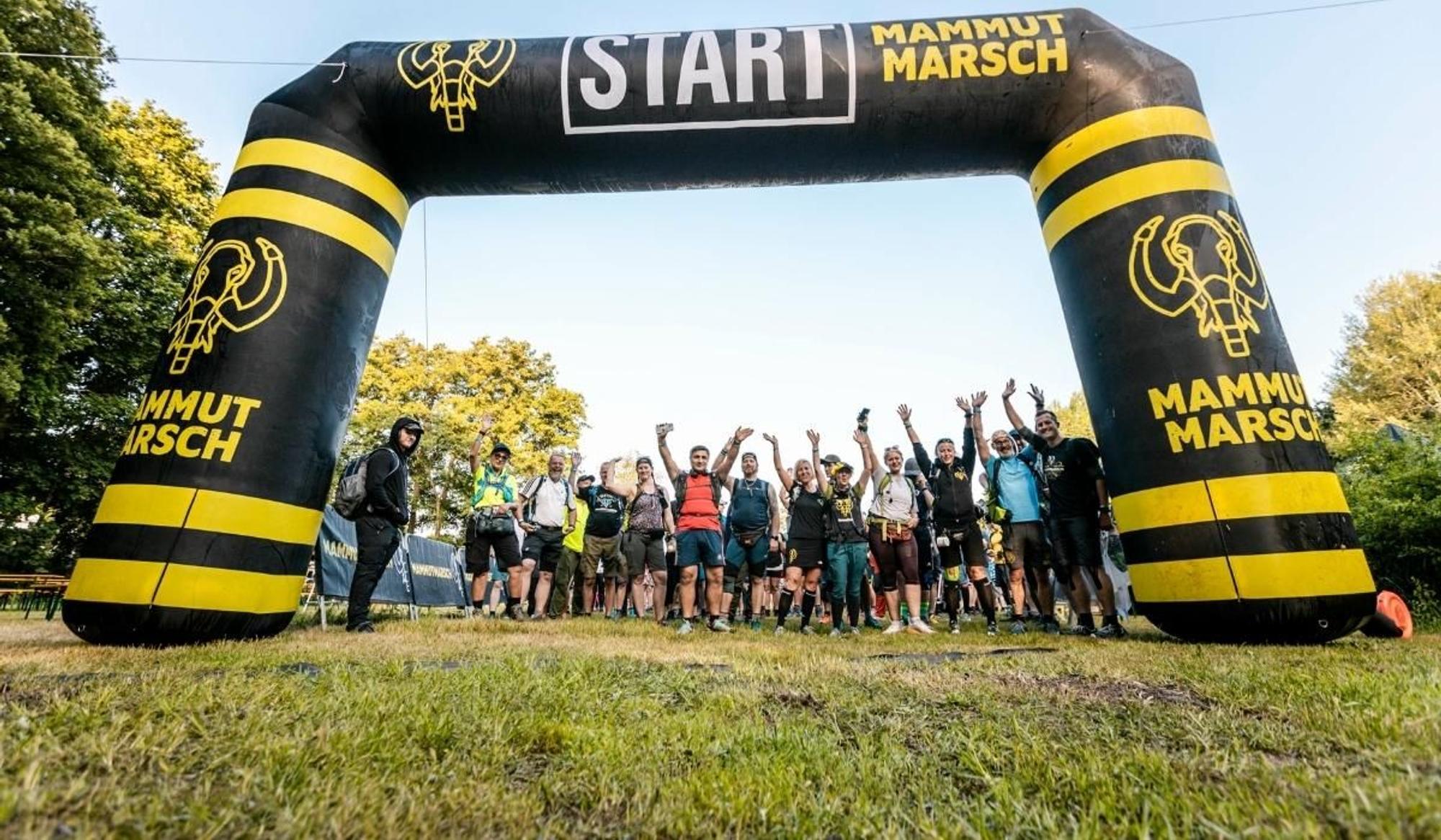 Gruppe fröhlicher Teilnehmer:innen steht winkend und jubelnd unter dem schwarz-gelben aufblasbaren Starttor des Mammutmarschs, auf dem „START" und das Mammutmarsch-Logo zu sehen sind – im Hintergrund grüne Bäume und blauer Himmel.