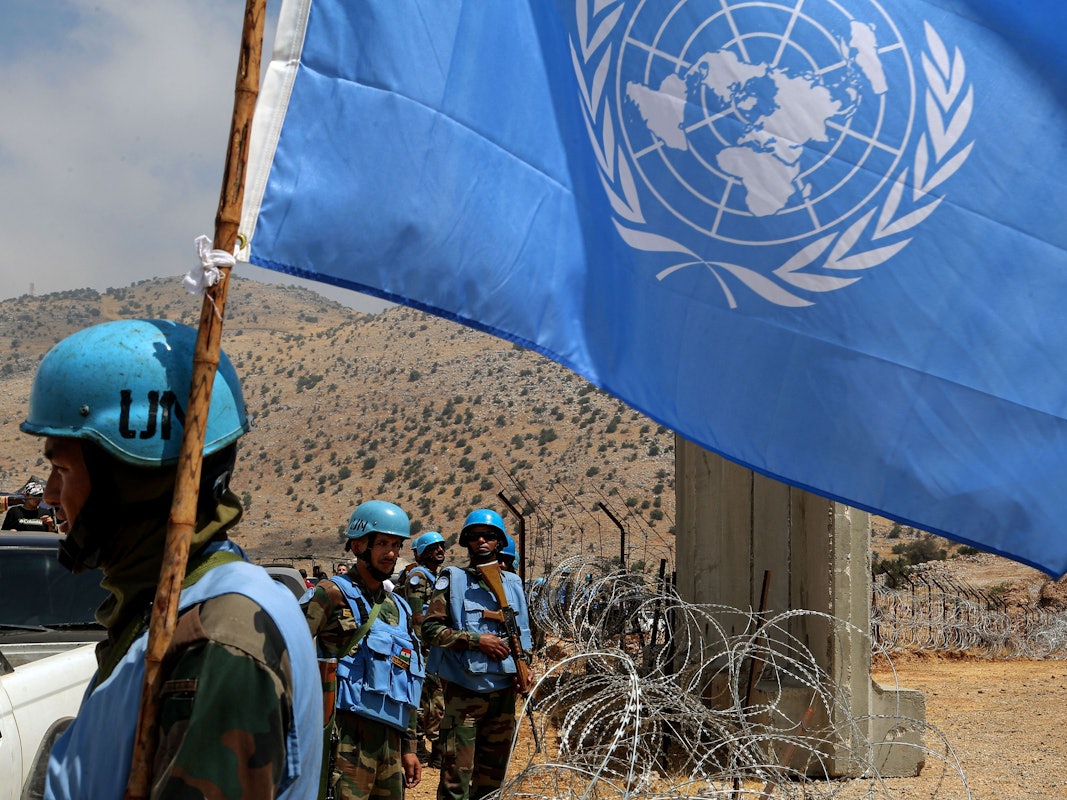 Mehrere Blauhelmsoldaten stehen an einem Stacheldrahtzaun, einer von ihnen trägt eine UN-Flagge.