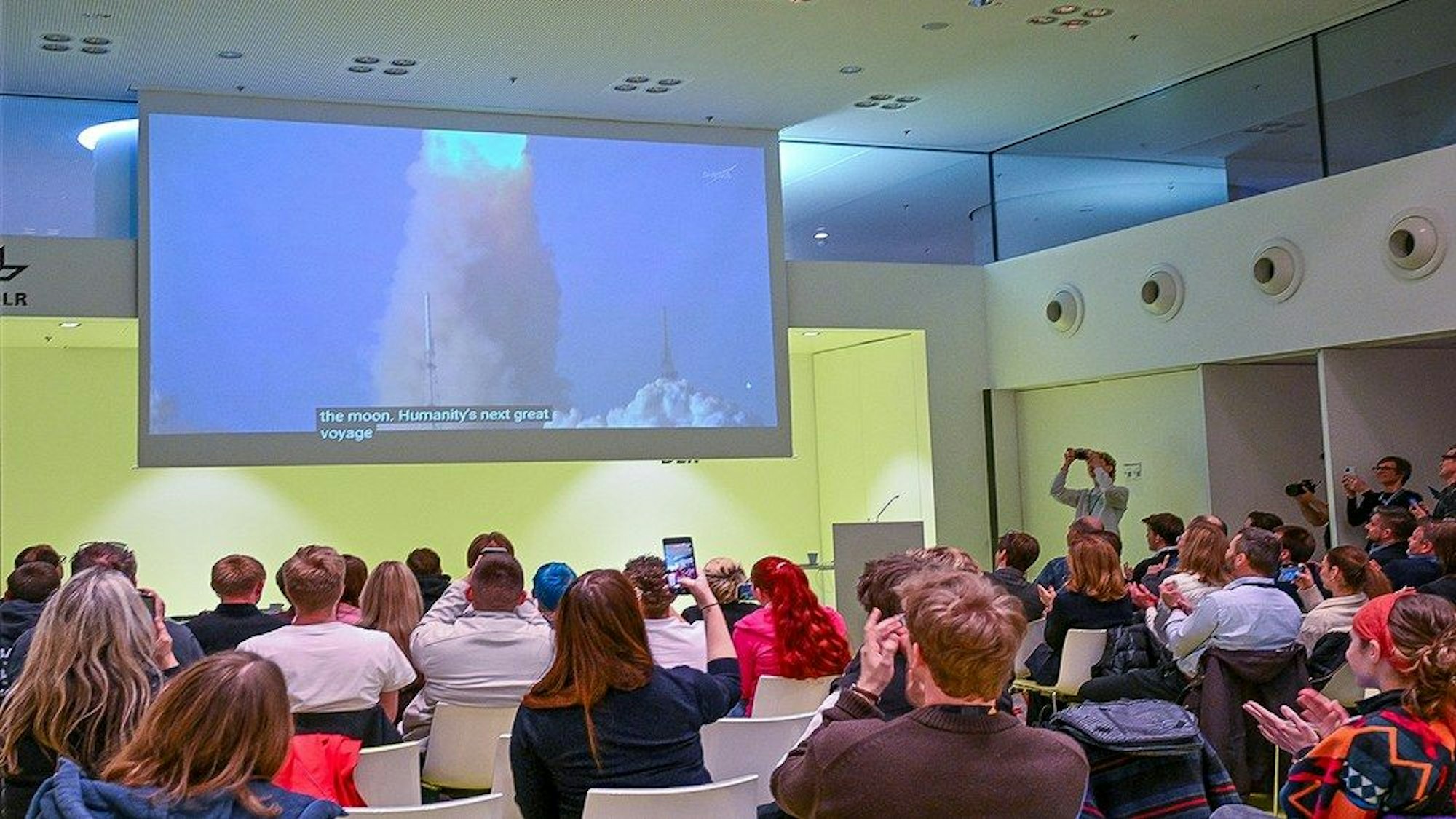 Menschen sitzen in einem Raum und gucken auf eine große Leinwand, auf der der Start einer Rakete gezeigt wird.