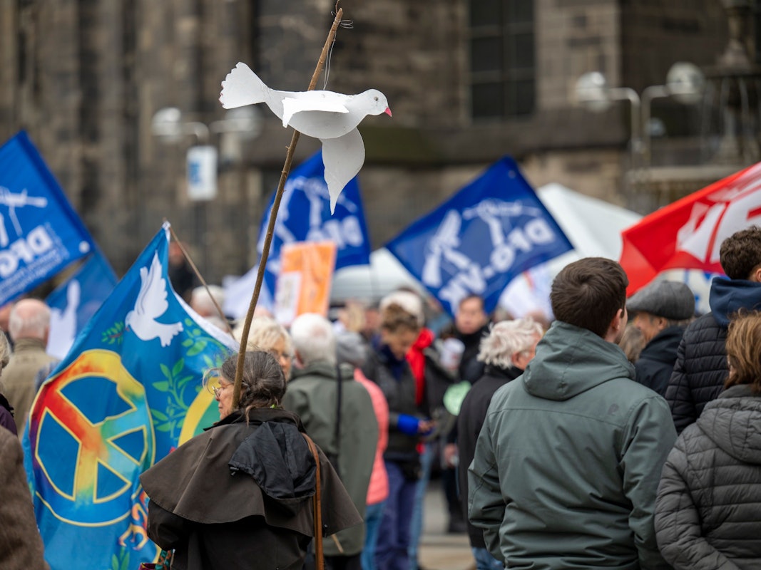 Menschen stehen mit Fahnen vor dem Kölner Dom. Eine Frau hält eine Friedenstaube auf Pappe an einem Stock in der Hand.