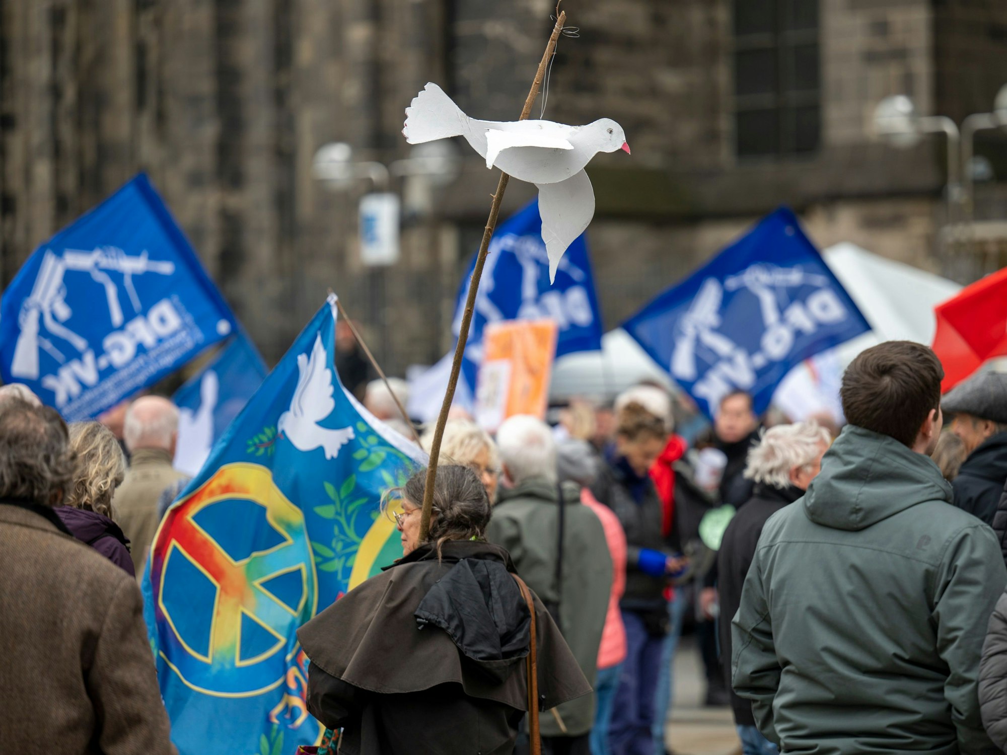 Menschen stehen mit Fahnen vor dem Kölner Dom. Eine Frau hält eine Friedenstaube auf Pappe an einem Stock in der Hand.