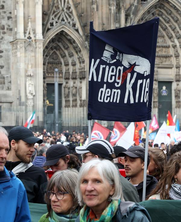 Menschen stehen gedrängt vor dem Kölner Dom, halten Fahnen und Plakate mit Aufschriften wie "Krieg dem Krieg" hoch.