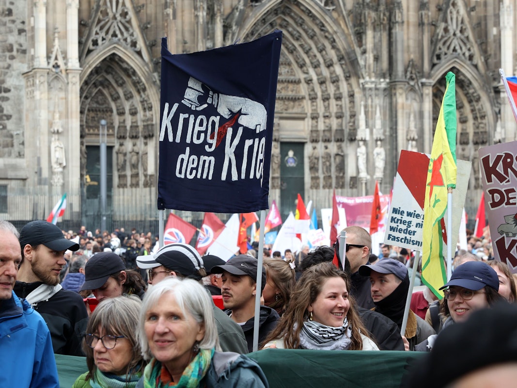 Menschen stehen gedrängt vor dem Kölner Dom, halten Fahnen und Plakate mit Aufschriften wie "Krieg dem Krieg" hoch.