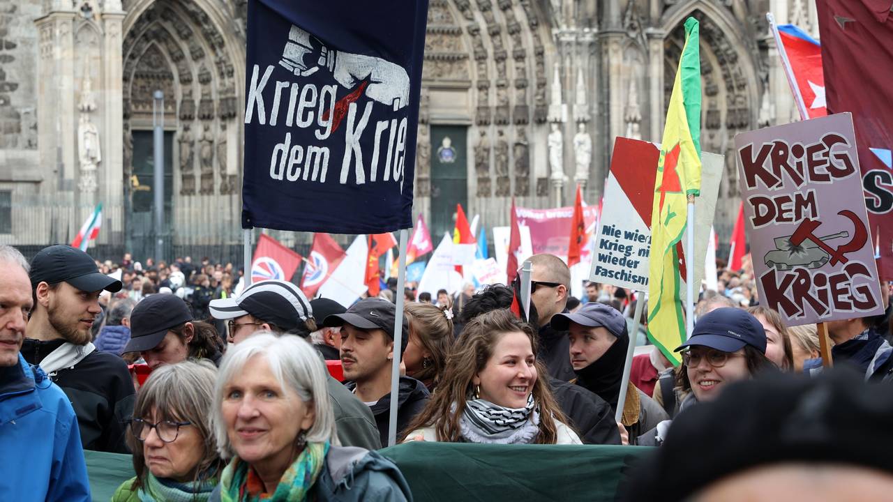 Menschen stehen gedrängt vor dem Kölner Dom, halten Fahnen und Plakate mit Aufschriften wie "Krieg dem Krieg" hoch.