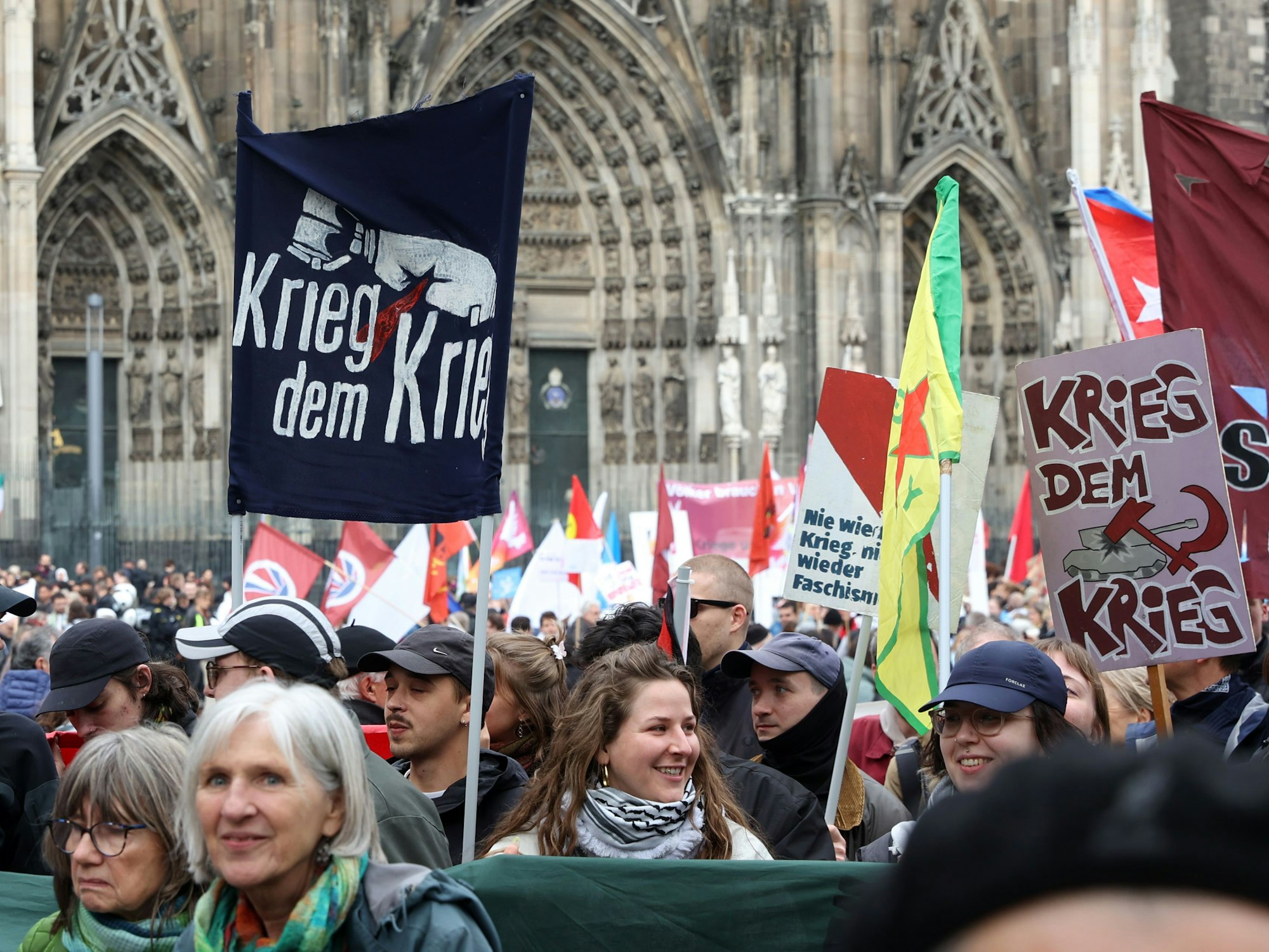 Menschen stehen gedrängt vor dem Kölner Dom, halten Fahnen und Plakate mit Aufschriften wie "Krieg dem Krieg" hoch.