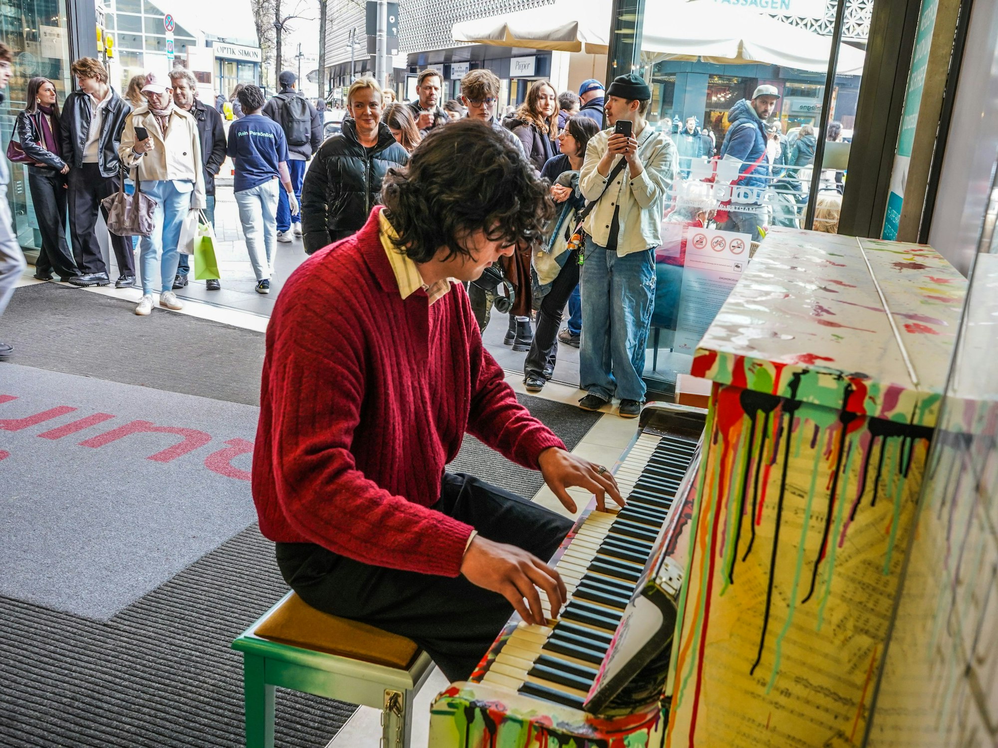 Ein junger Mann spielt an einem öffentlichen Klavier, hinter ihm stehen Leute, teilweise mit Handys.