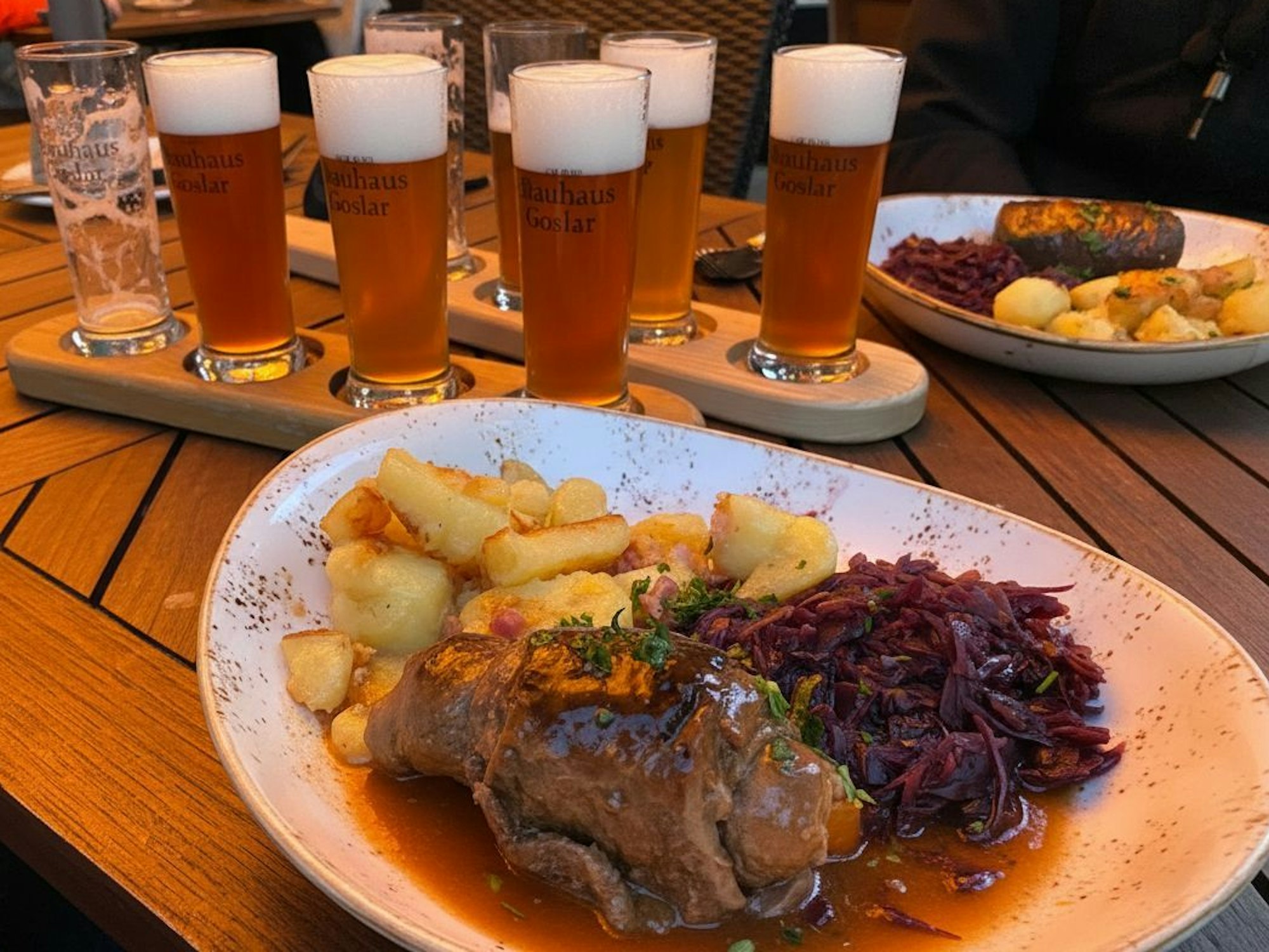 Ein Teller mit Rouladen, Rotkraut und Schupfnudeln, dahinter stehen Holzbretter mit gefüllten Biergläsern