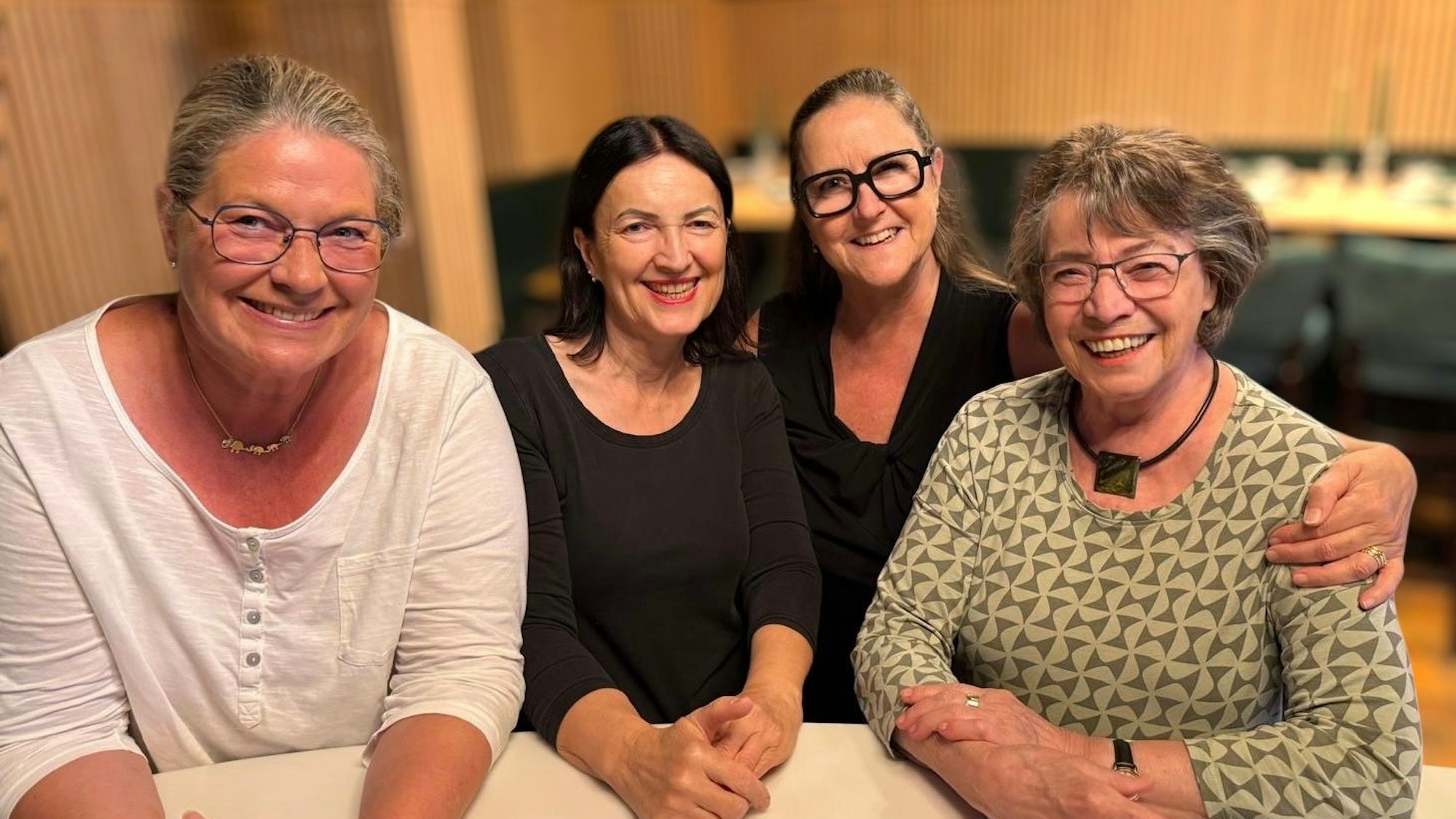 Vier Frauen mittleren Alters sitzen nebeneinander an einem Tisch und freuen sich.