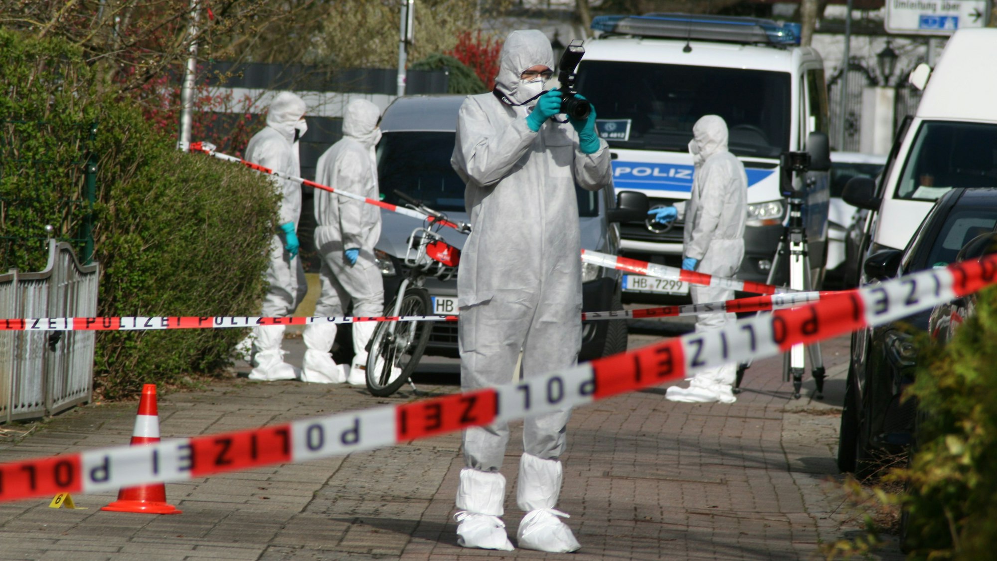 Vier Personen ind Schutzanzügen sind an dem Tatort, der mit Polizeiflatterband abgesperrt ist.