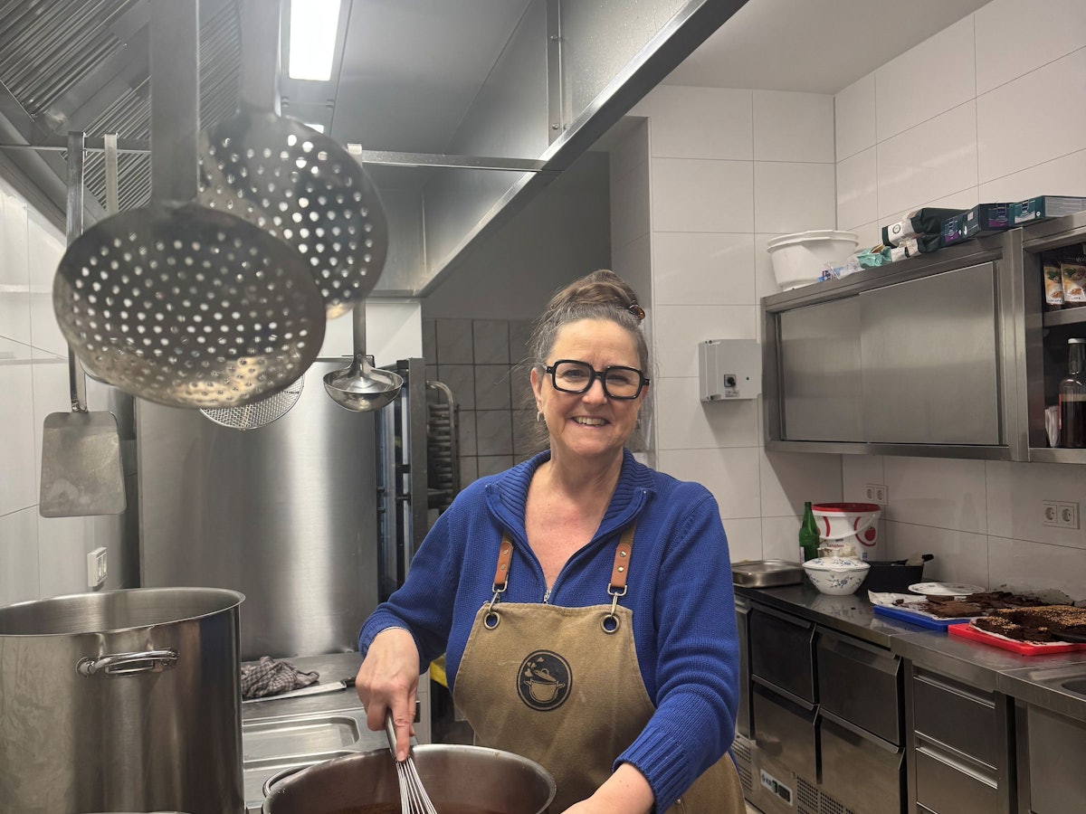 Eine Dame mittleren Alters mit markanter Brille, Dutt und Küchenschürze steht an einem Herd und rührt mit einem großen Schneebesen in einem Topf.