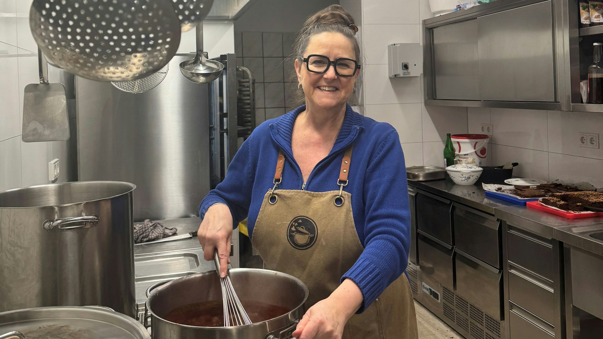 Eine Dame mittleren Alters mit markanter Brille, Dutt und Küchenschürze steht an einem Herd und rührt mit einem großen Schneebesen in einem Topf.