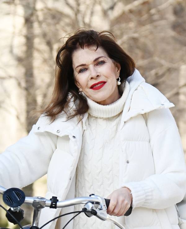 Eine ältere Frau mit langen dunklen Haaren und weißer Daunenjacke schieb ein Fahrrad und lächelt mit knallrot geschminktem Mund in die Kamera.