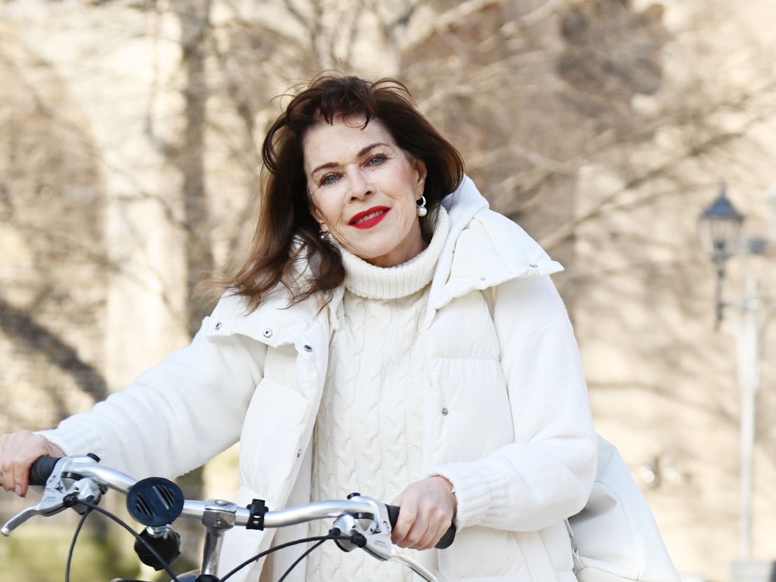 Eine ältere Frau mit langen dunklen Haaren und weißer Daunenjacke schieb ein Fahrrad und lächelt mit knallrot geschminktem Mund in die Kamera.