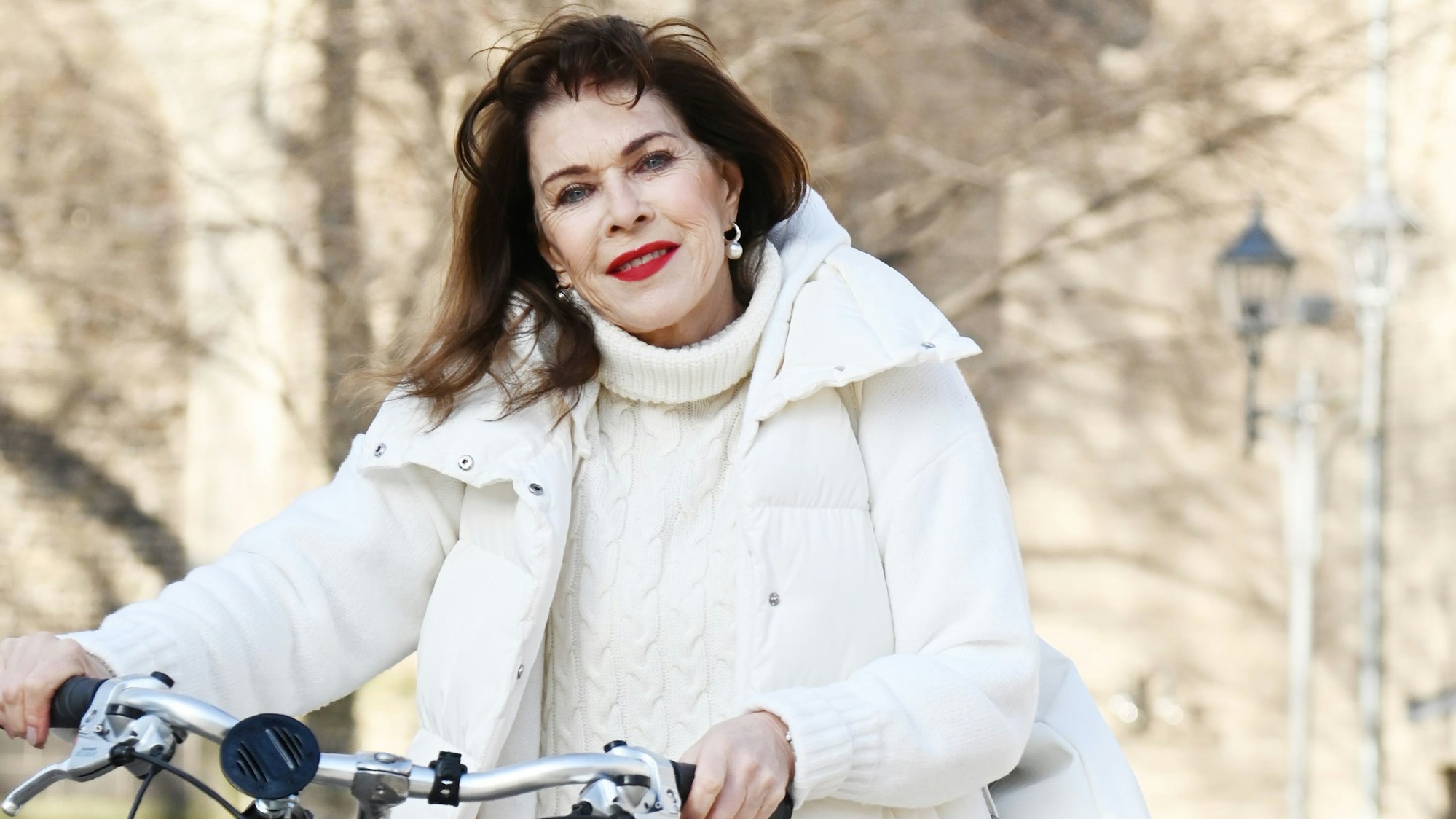 Eine ältere Frau mit langen dunklen Haaren und weißer Daunenjacke schieb ein Fahrrad und lächelt mit knallrot geschminktem Mund in die Kamera.