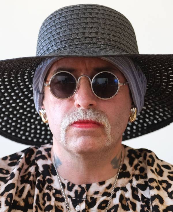 Eine Frau trägt Hut, Brille und Schnurbart, ihre Lippen sind rot geschminkt.