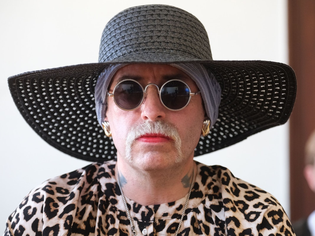 Eine Frau trägt Hut, Brille und Schnurbart, ihre Lippen sind rot geschminkt.